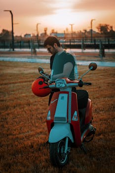 A man stands beside his red scooter, enjoying the sunset outdoors.