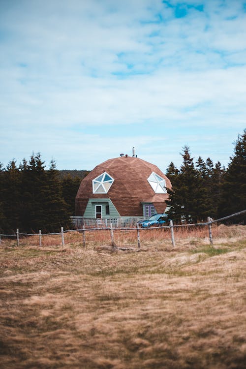 Exterior of creative contemporary residential house with poly angular piled roof of brown color and star shaped window located on grassy valley amidst firs and rural fence