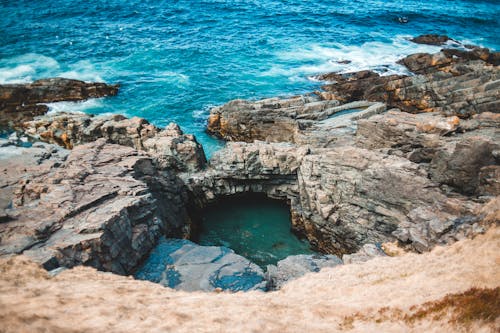 From above of amazing waving turquoise ocean with rocky cliffs and grotto on sunny day