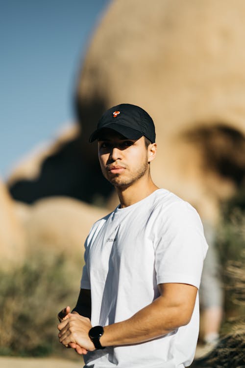 Confident young ethnic male in white t shirt and cap standing ear rocky cliff on sunny day and looking at camera