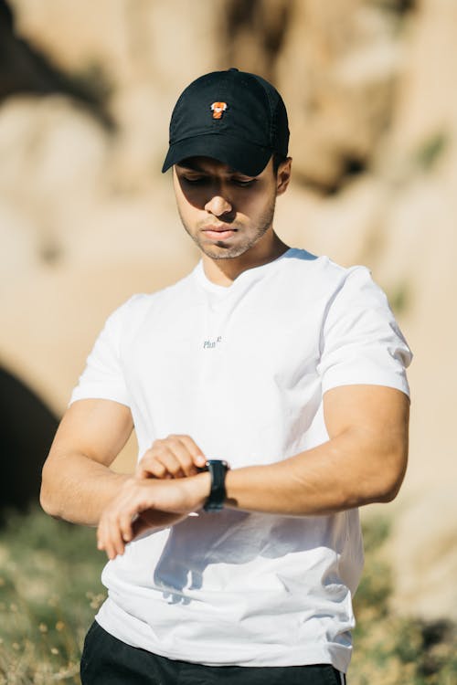 Free Serious young ethnic guy in white t shirt and cap checking time on wristwatch while waiting for meeting in park on sunny day Stock Photo