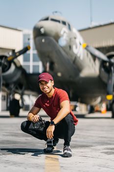 Full body of happy young ethnic male in stylish outfit sitting on haunches and looking at camera near aircraft in aerodrome