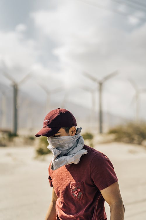 Anonymous young man with bandana on face standing near windmills in desert valley