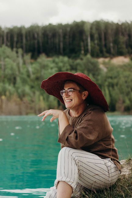 A joyful senior woman wearing a sun hat sits by a serene lake, surrounded by nature.