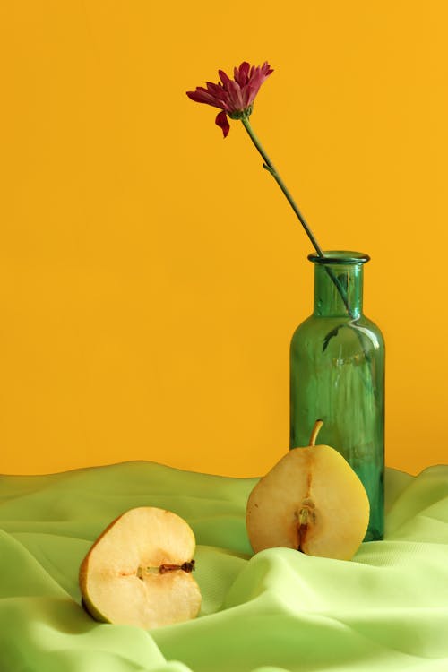 Ripe fresh sweet halved pear placed on textile near green glass vase with delicate flower against yellow background