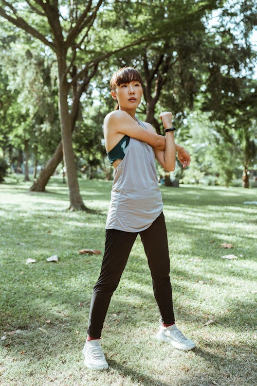 Full length focused adult Asian female athlete in sportswear standing in park while warming up muscles before training alone