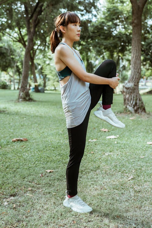 Free Side view full length of concentrated young Asian sportswoman in activewear standing in sunny green park and stretching legs while warming up and looking away Stock Photo