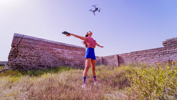 Full body stylish female in shorts and top standing with arms outstretched under flying drone on grassy meadow near ruined brick wall