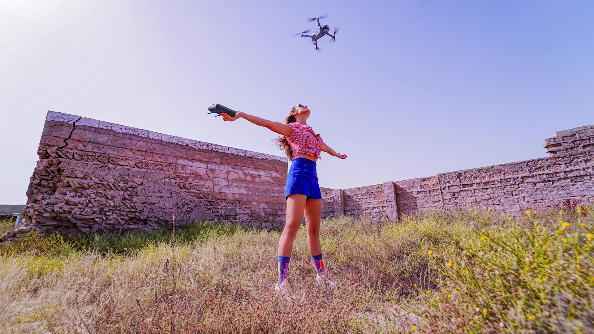 Young woman controlling drone in an open meadow with an old stone wall.