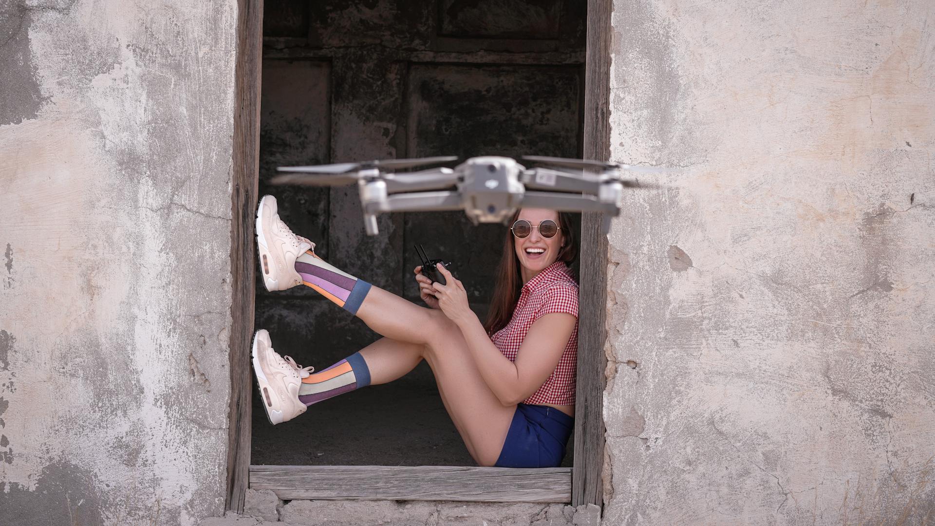 Side view full body excited female in casual wear sitting at remaining hut entrance and operating modern drone