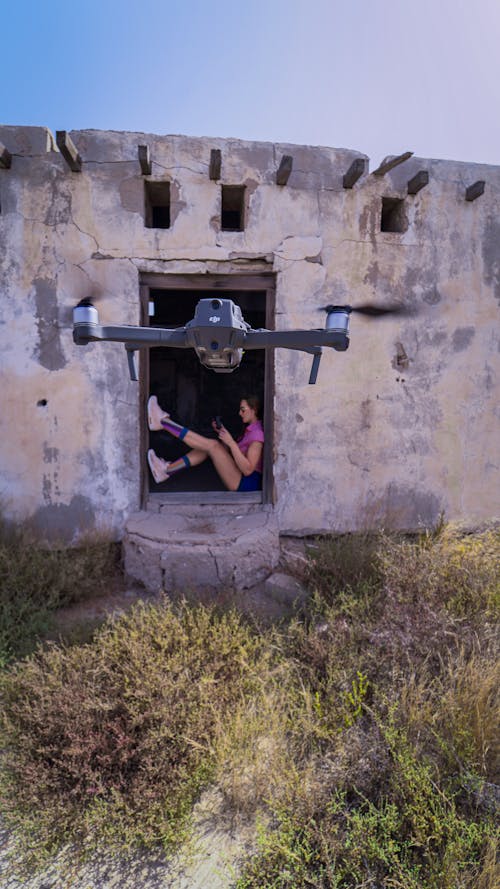 Side view full length stylish female resting at abandoned hut with legs raised and operating contemporary drone