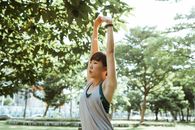 一位穿着运动服的亚洲女士，在阳光明媚的夏季公园里，正冷静而积极地做着伸展手臂的练习