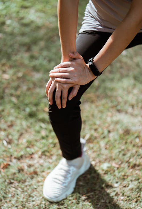 Crop anonymous slim sportswoman in black leggings and white sneakers stretching legs before jogging while warming up on green meadow in sunlight