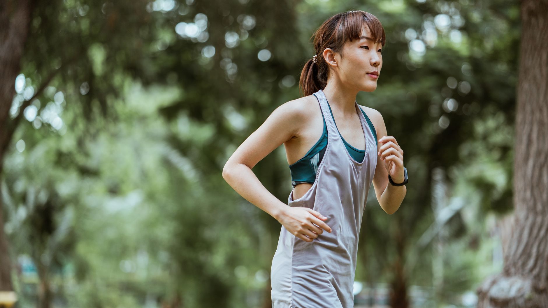 Side view of fit young Asian female jogger wearing gray sports top running along path in sunny summer park