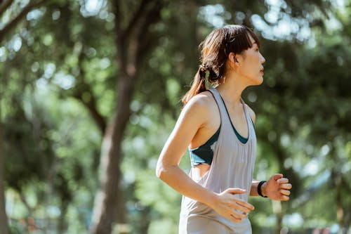 Side view of determined focused Asian female runner in sportswear jogging in green lush park on sunny morning