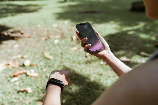 From above crop unrecognizable female wearing fitness tracker browsing modern mobile phone with blank screen while resting in green park