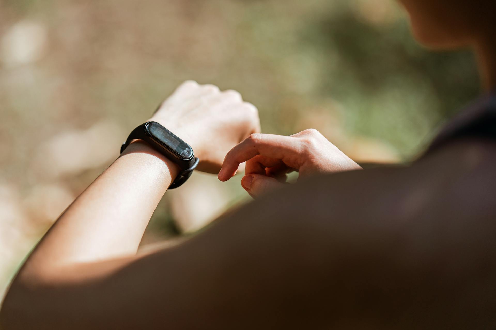 From behind crop anonymous sportswoman checking notifications on  modern fitness tracker while training against blurred nature background on sunny weather