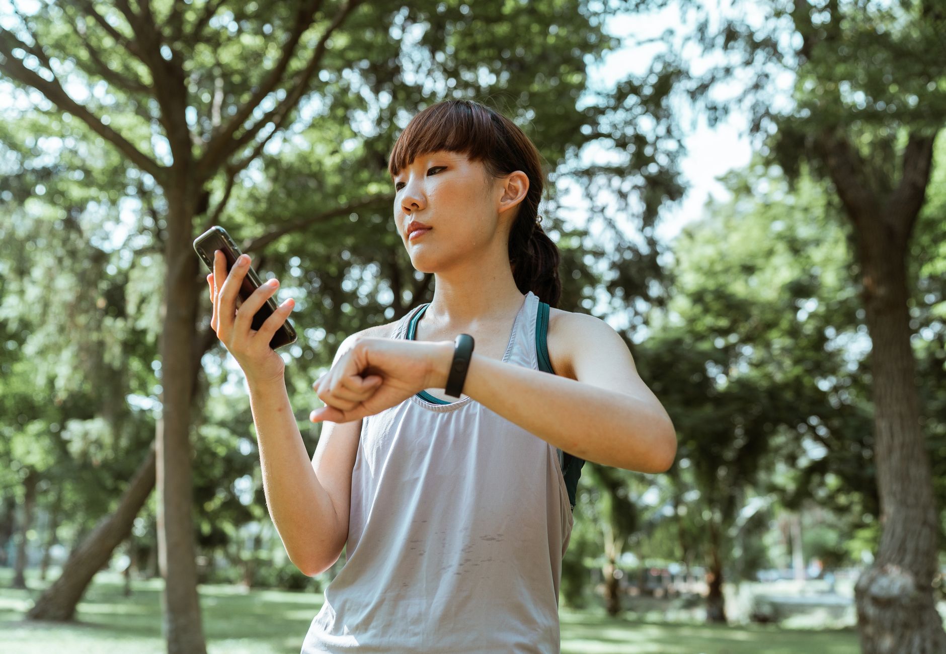 Positive concentrated young fit Asian sportswoman in activewear browsing smartphone and checking fitness bracelet while training in green sunny park