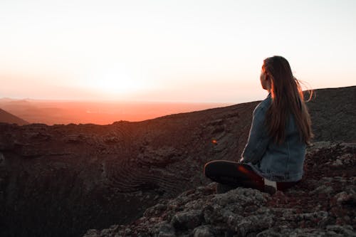 Woman in Blue Denim Jacket Sitting on Rock during Sunset