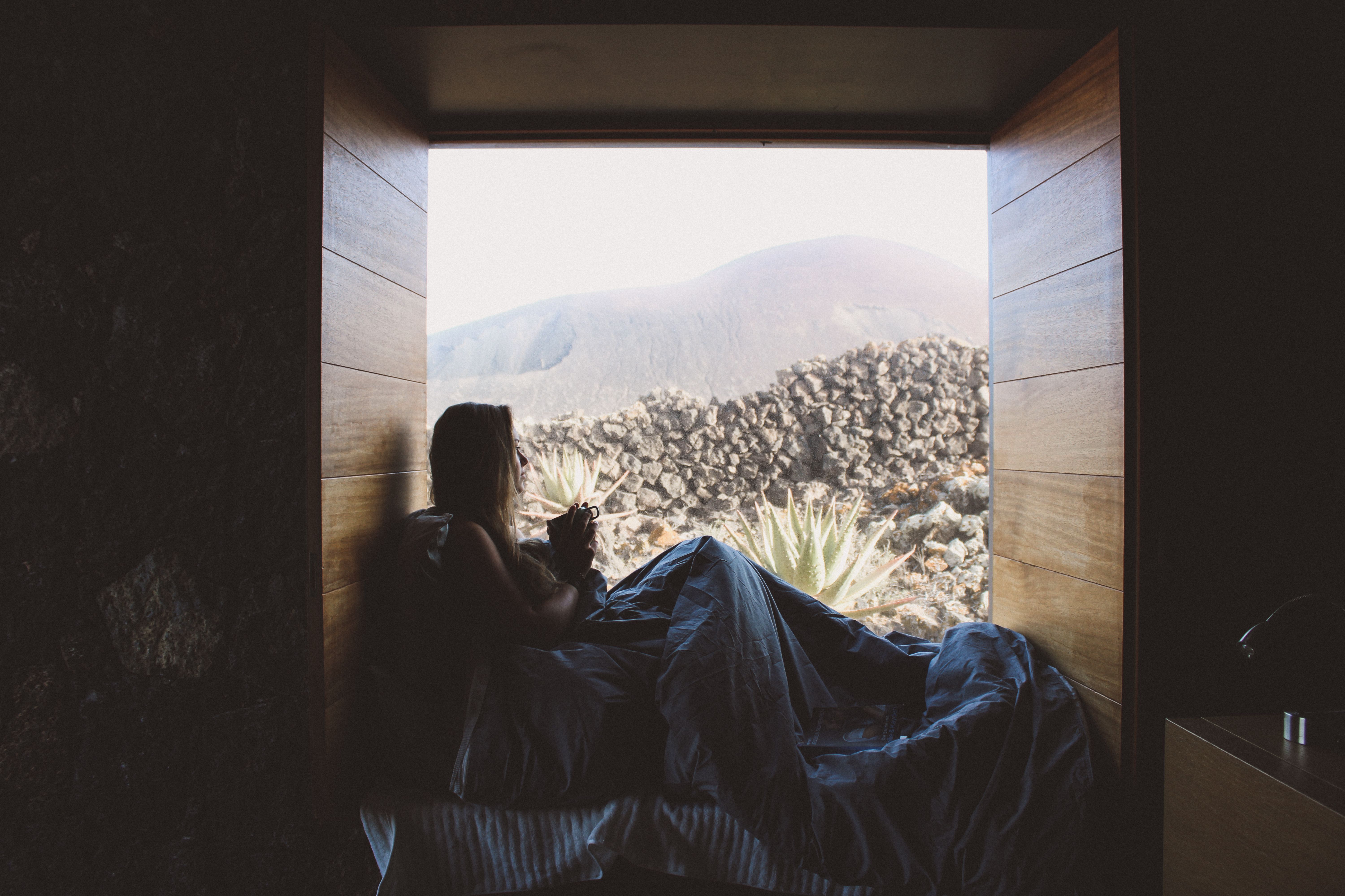 Silhouette of a woman in bed enjoying a mountain view through a rustic window.