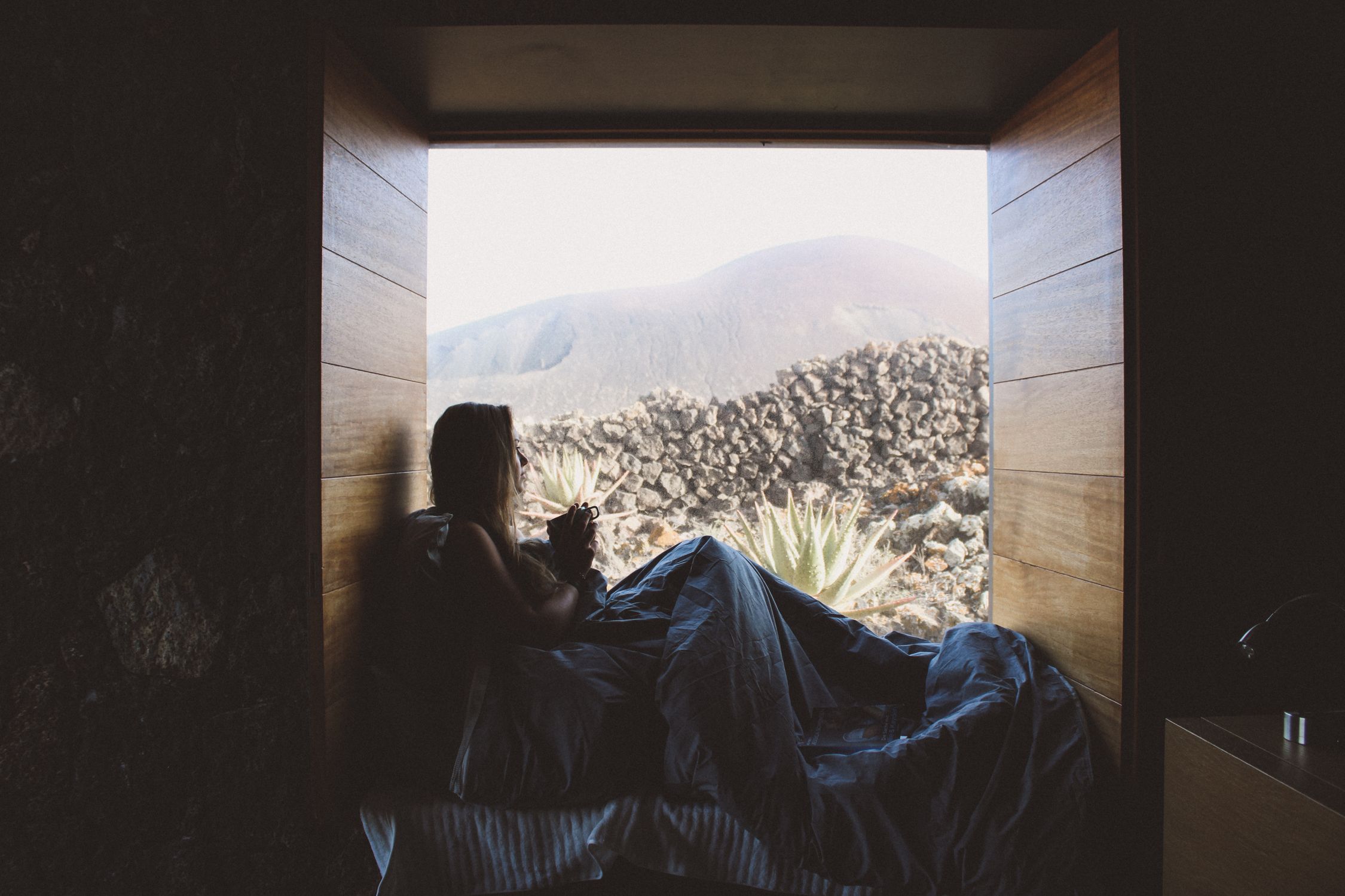 Silhouette of a woman in bed enjoying a mountain view through a rustic window.
