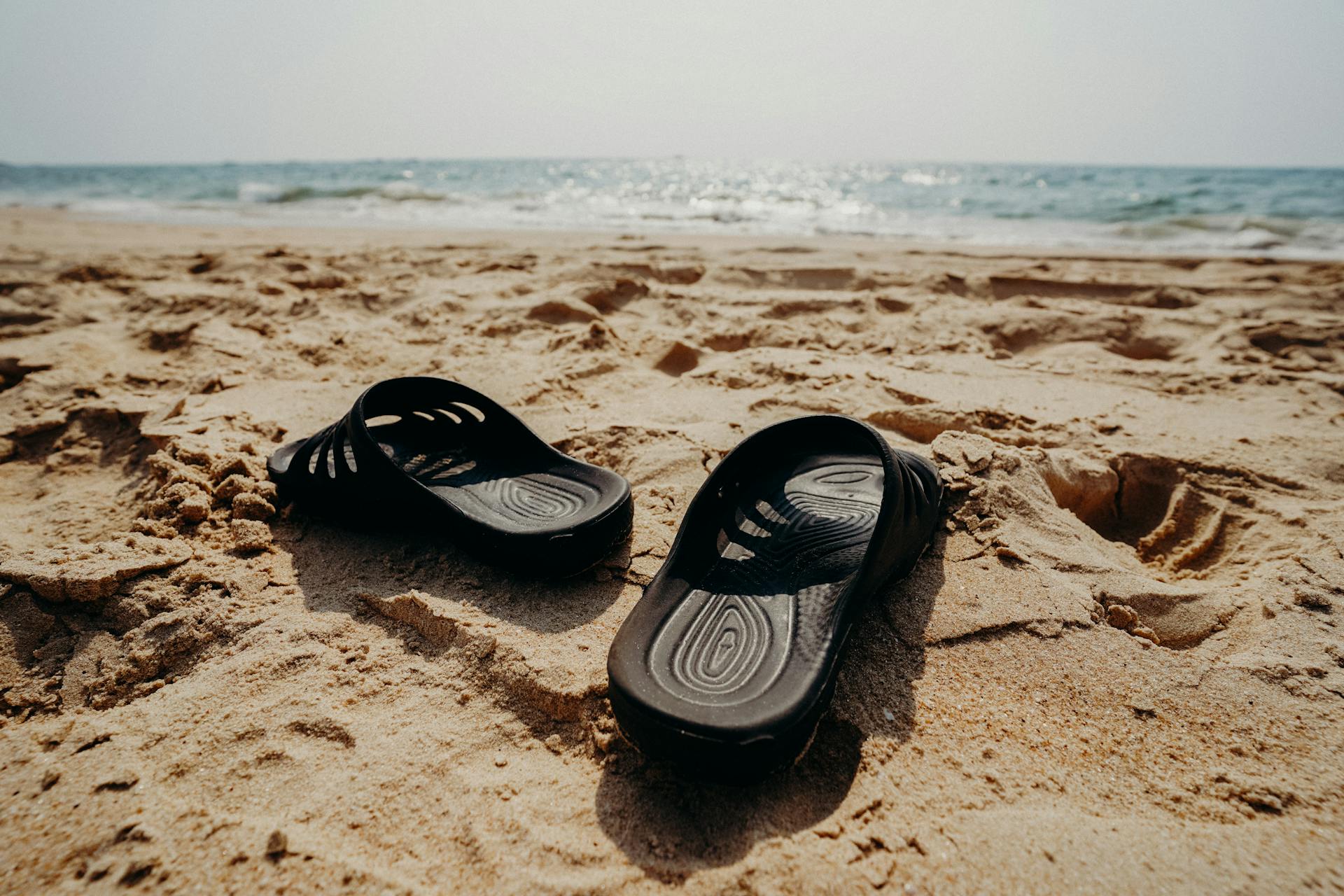 Casual sandals resting on a sunlit beach with ocean waves in the background, ideal for summer vacation themes.