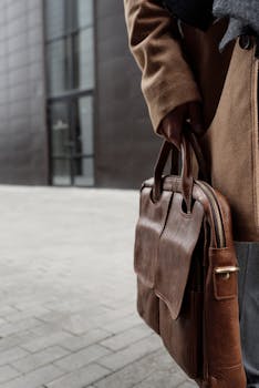 Person holding a stylish brown leather briefcase outdoors in a modern urban setting.
