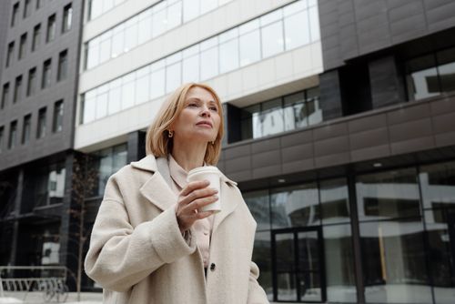 Free Elegant woman in beige coat holding coffee cup outside modern building. Stock Photo