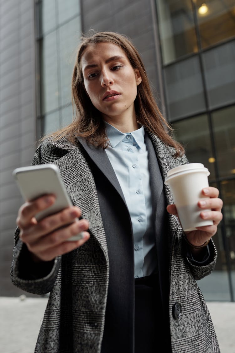 A Woman Browsing Her Smartphone While Holding A Cup Of Coffee