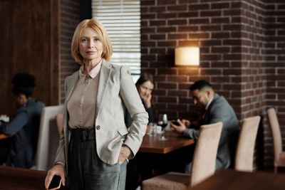 Mature businesswoman in gray blazer standing confidently in a stylish café interior.