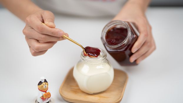 From above crop faceless person adding berry jam to plain organic yogurt in glass jar placed on wooden saucer near tiny cat statuette