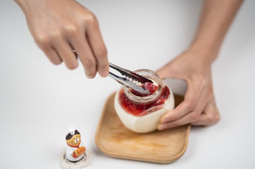 From above crop anonymous person adding organic ripe raspberries into jar with homemade tasty yogurt placed on white table near funny cat statuette