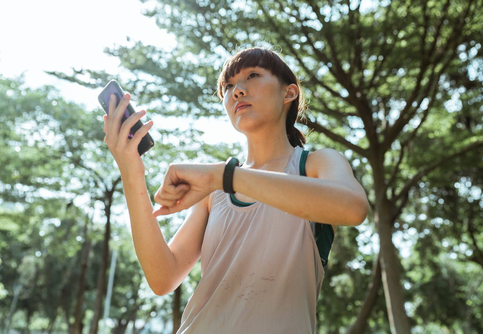 From below ethnic Asian female wearing sport clothes standing in park and connecting smartphone and smart watch during break in sunny day