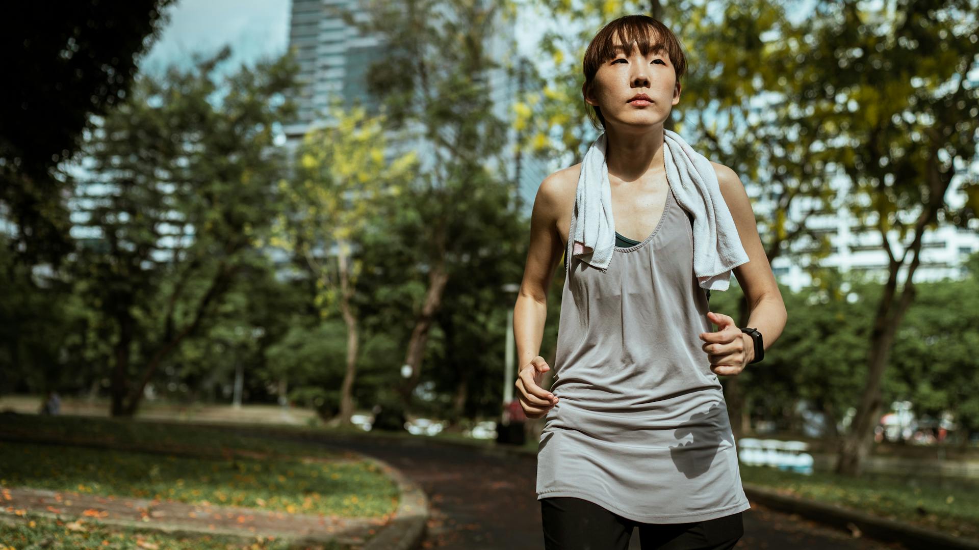 Serious Asian female runner wearing sportswear looking away while jogging on asphalt road in sunny day in park during training