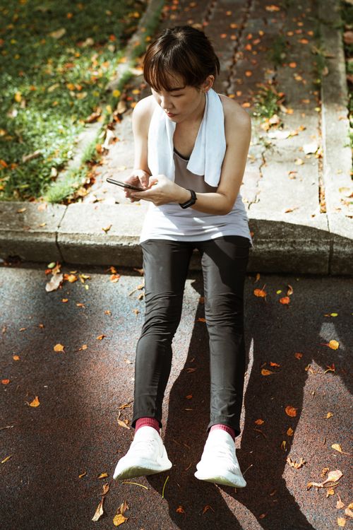 Free From above of ethnic Asian serious woman wearing sport clothes sitting on border and browsing smartphone while resting after training in park Stock Photo