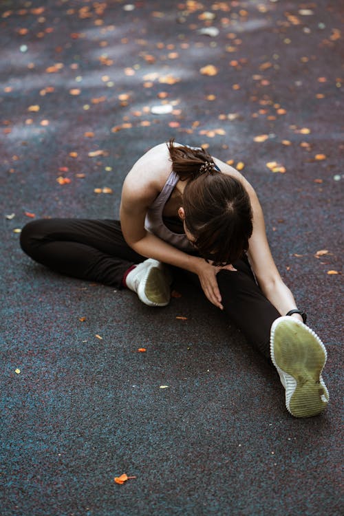 Anonymous flexible female in sportswear and sneakers sitting on pavement and bending forward while stretching on sports ground