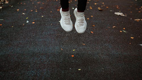 Feet of faceless person in white sneakers jumping above dark surface of sports ground