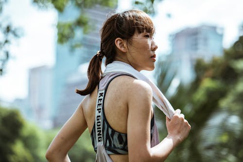 Side view of ethnic female in activewear with towel over neck looking away while resting after gymnastic in sunny day