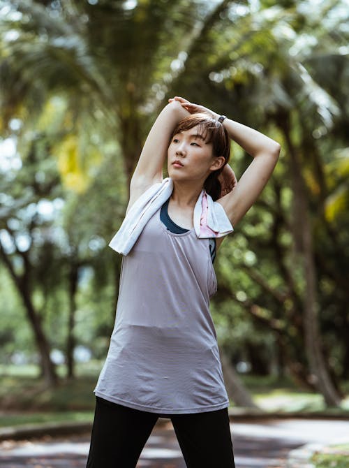 Pensive ethnic flexible female in sportswear stretching hands and looking away while warming up in park