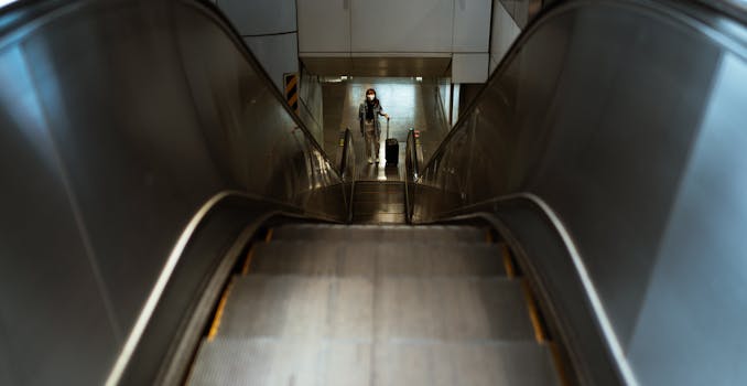 A lone traveler wearing a face mask stands on an escalator with luggage, emphasizing themes of travel and pandemic safety.