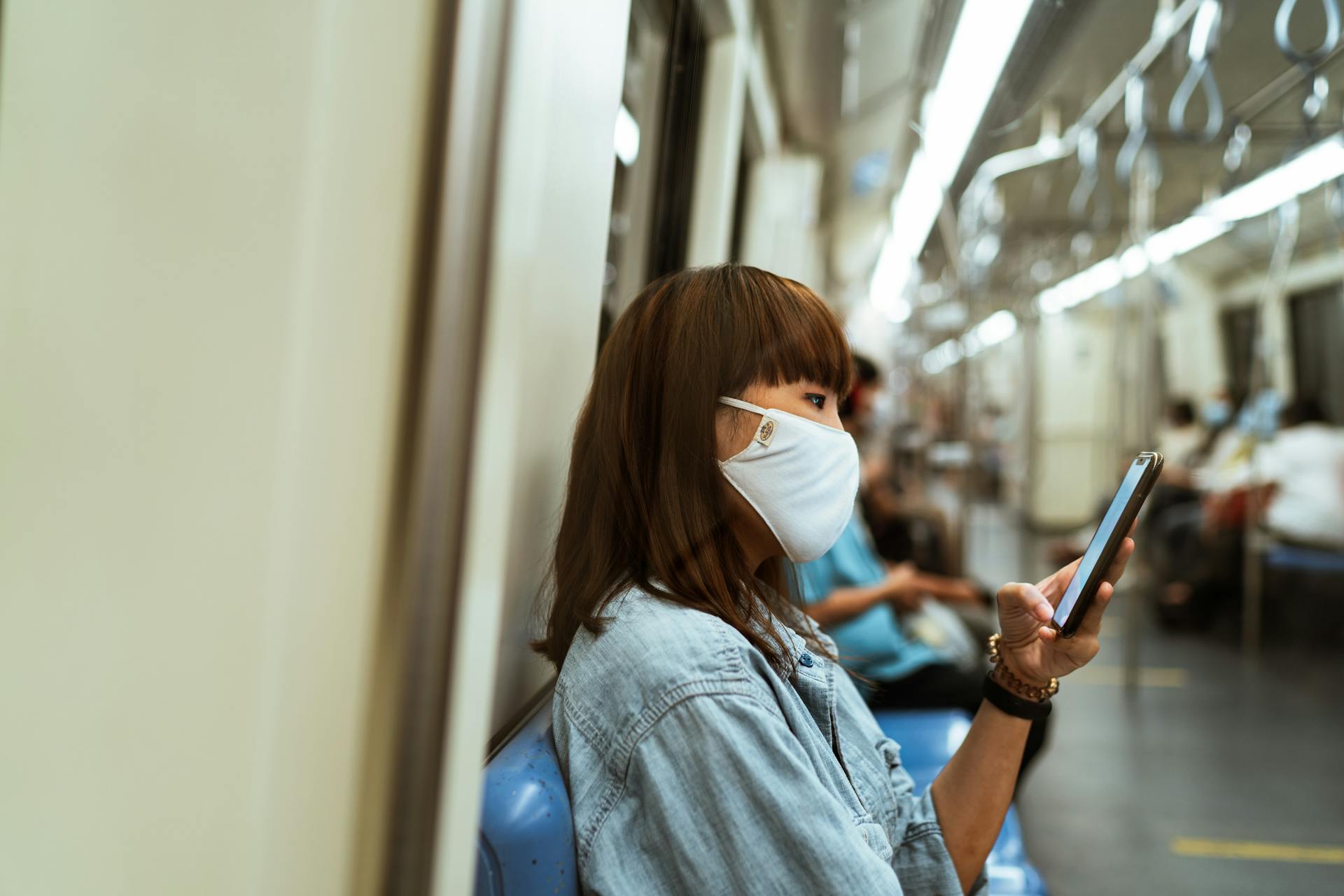 Asian woman wearing face mask using smartphone in subway, emphasizing public transport during pandemic.