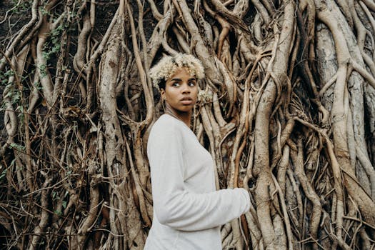 A young woman with curly hair stands in front of intricately twisted tree roots, showcasing a natural backdrop.