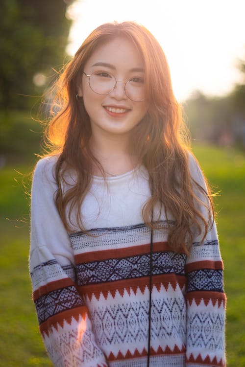 Charming young Asian female in casual sweater standing on abundant lush meadow and looking at camera with smile