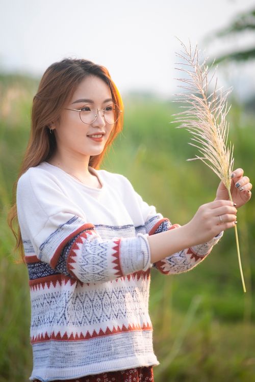 Free Optimistic young Asian female in casual outfit standing on lush meadow and looking at cereal spike in hands Stock Photo