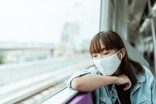 一位亚洲妇女白天在火车上戴着口罩和耳机休息