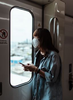 一名亚洲女子戴着口罩，手持智能手机，透过火车车窗向外凝视