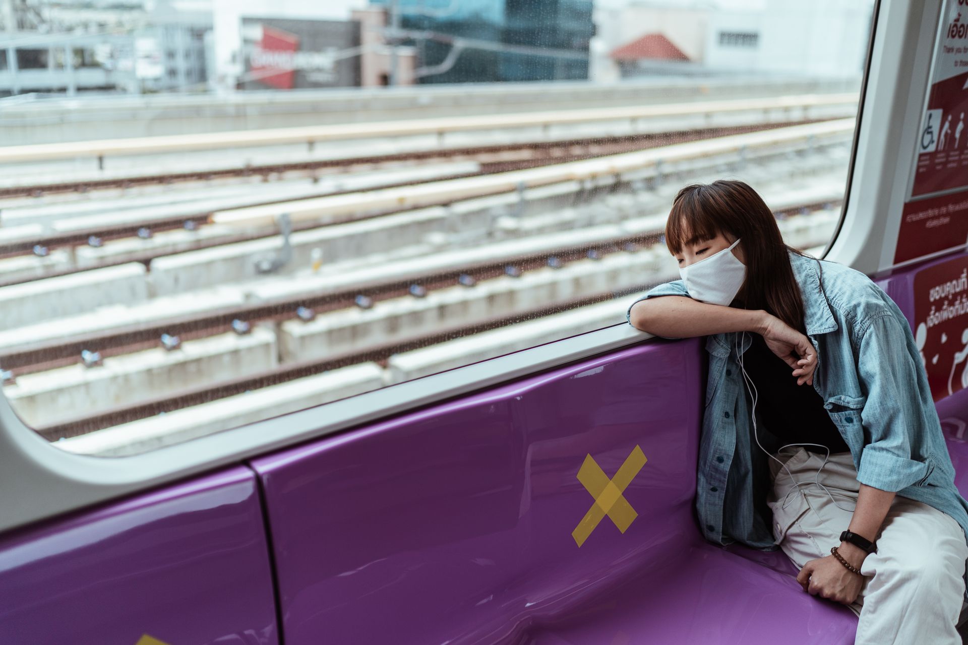 A woman wearing a face mask rests on a subway train seat with a view of the tracks outside.