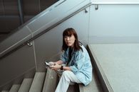 一位穿着休闲装、佩戴智能手表的严肃亚洲女性，正坐在大楼楼梯上，从侧上方看着摄像机，使用手机社交媒体