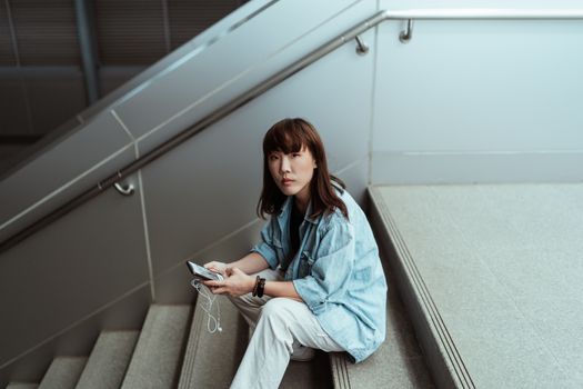 一位穿着休闲装、佩戴智能手表的严肃亚洲女性，正坐在大楼楼梯上，从侧上方看着摄像机，使用手机社交媒体
