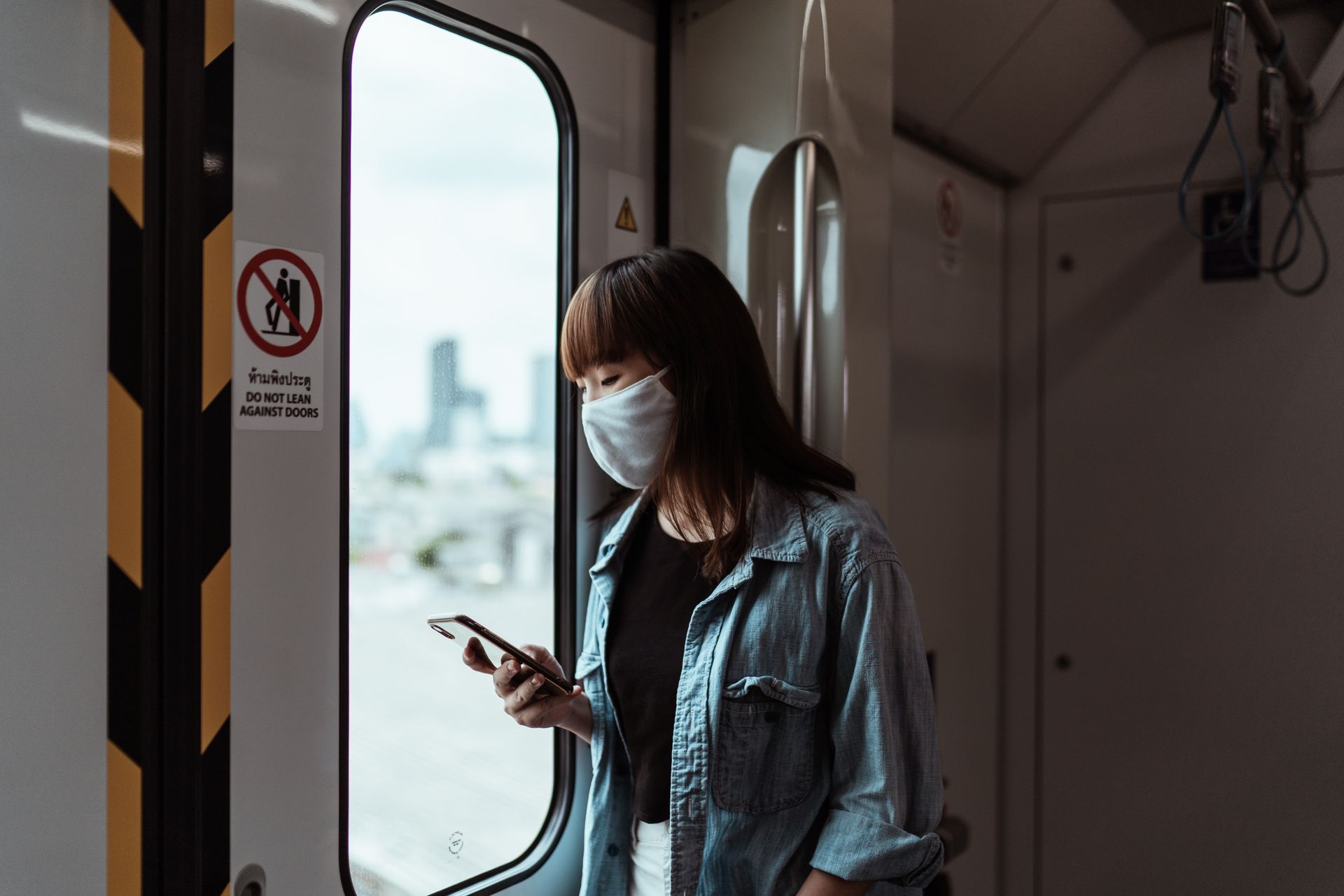 Asian woman wearing a face mask, using a smartphone on a train during the day.